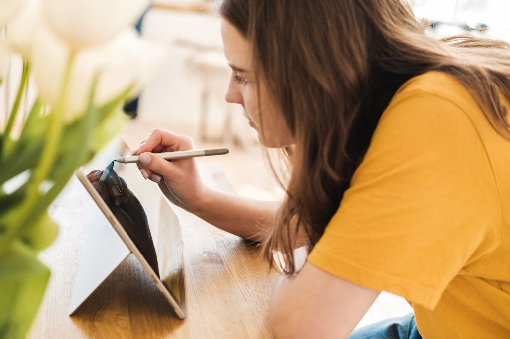 Woman using a stylus on a tablet at a bright table with flowers nearby